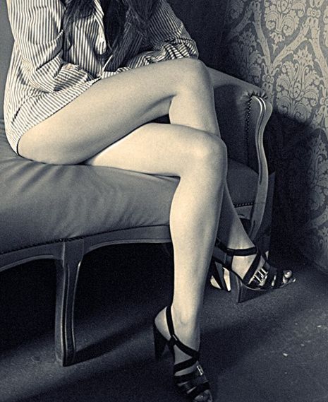 A seated woman with crossed legs in strappy heels, on an upholstered chair.