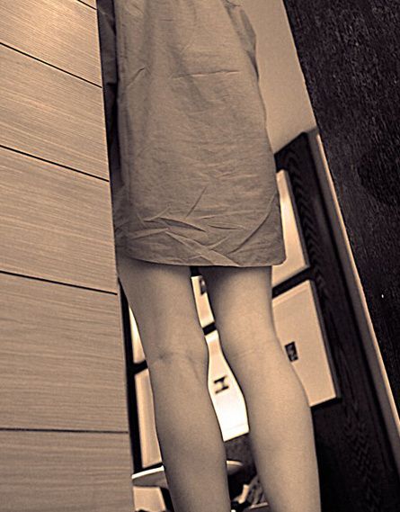 Bare legs below a short skirt, standing by a wooden wall, in sepia.