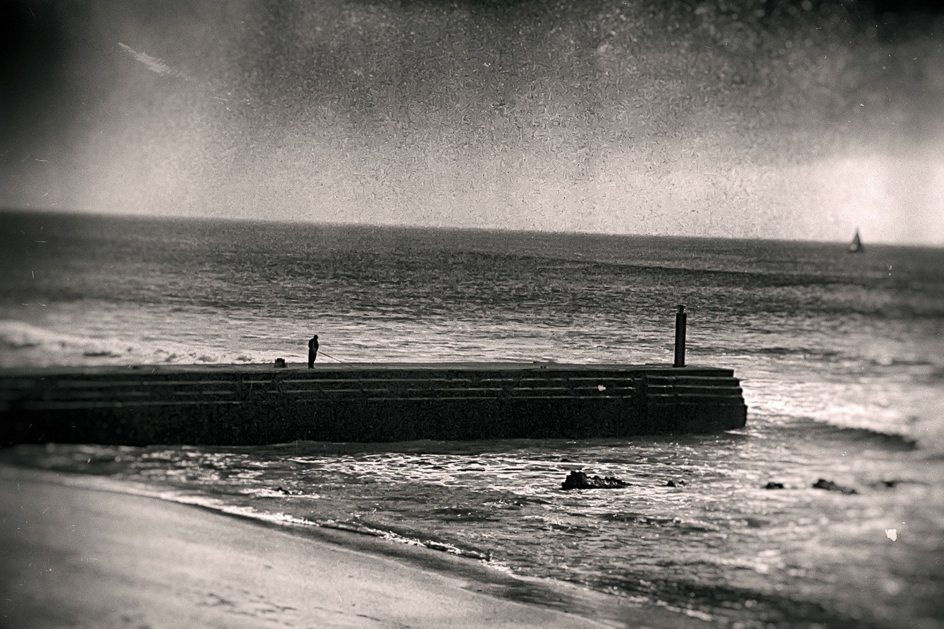 Black-and-white coastline: a stone jetty reaching into a rough sea, two small figures at its end.