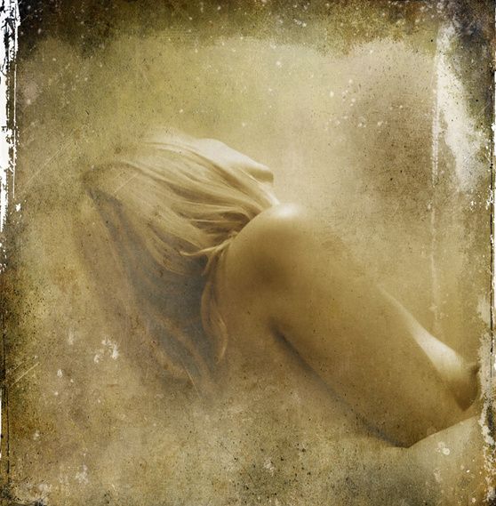 A nude woman seen from behind, blonde hair falling forward, in a hazy golden light.