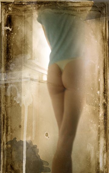 A woman in a light top seen from behind by a bright window, in a worn sepia frame.