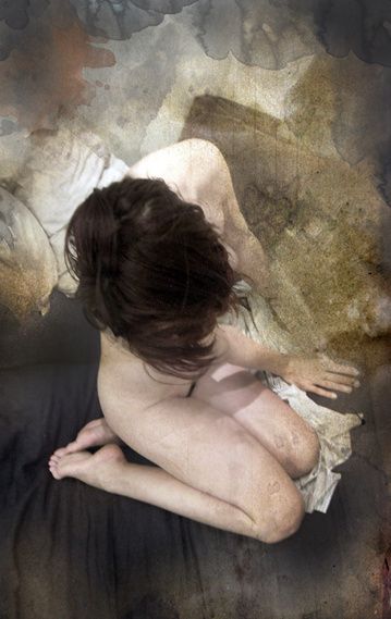 A dark-haired woman sitting curled on a pale sheet, seen from above.