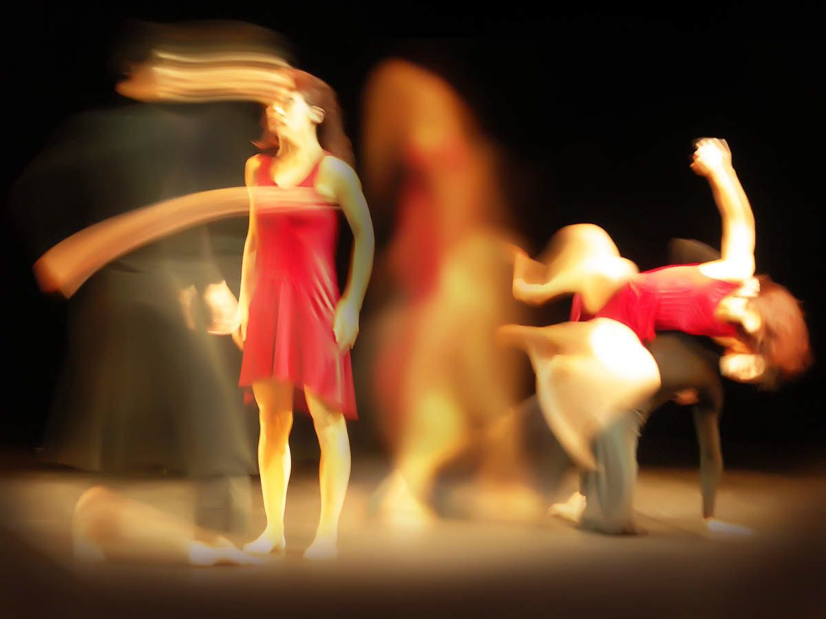 A dancer in a red dress standing still while others whirl around her in blurred light.