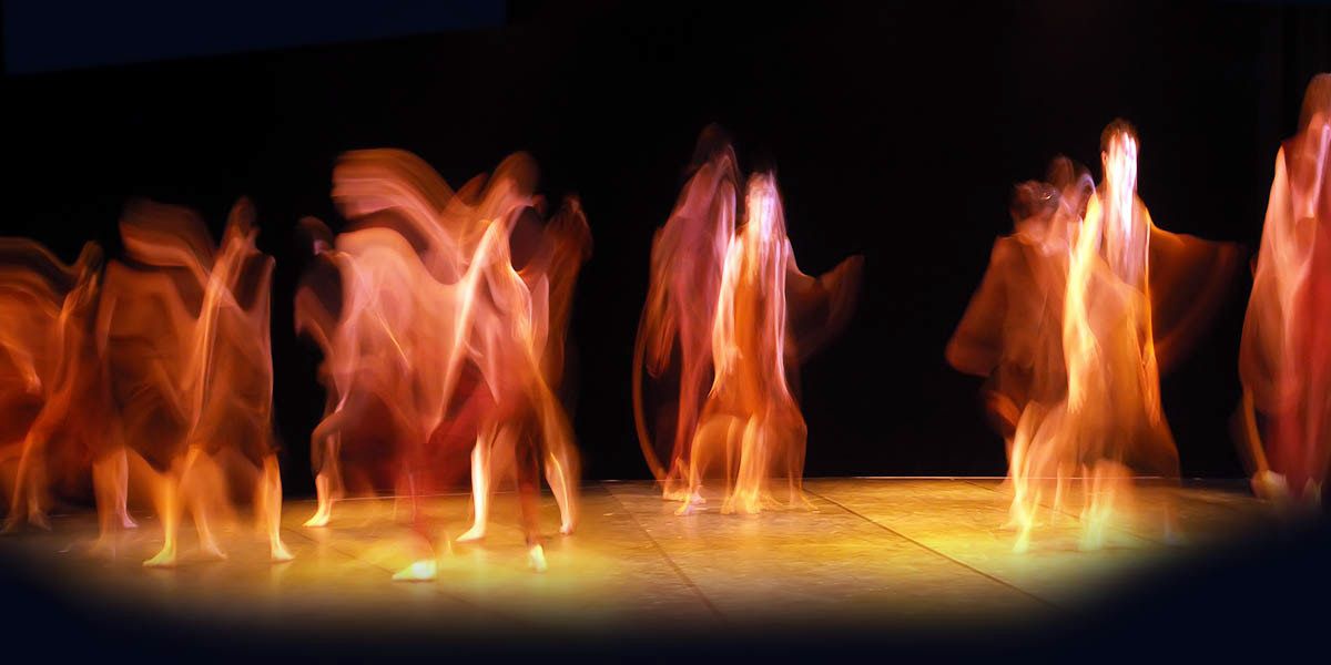 Panoramic stage: a line of dancers dissolving into streaks of amber light against black.