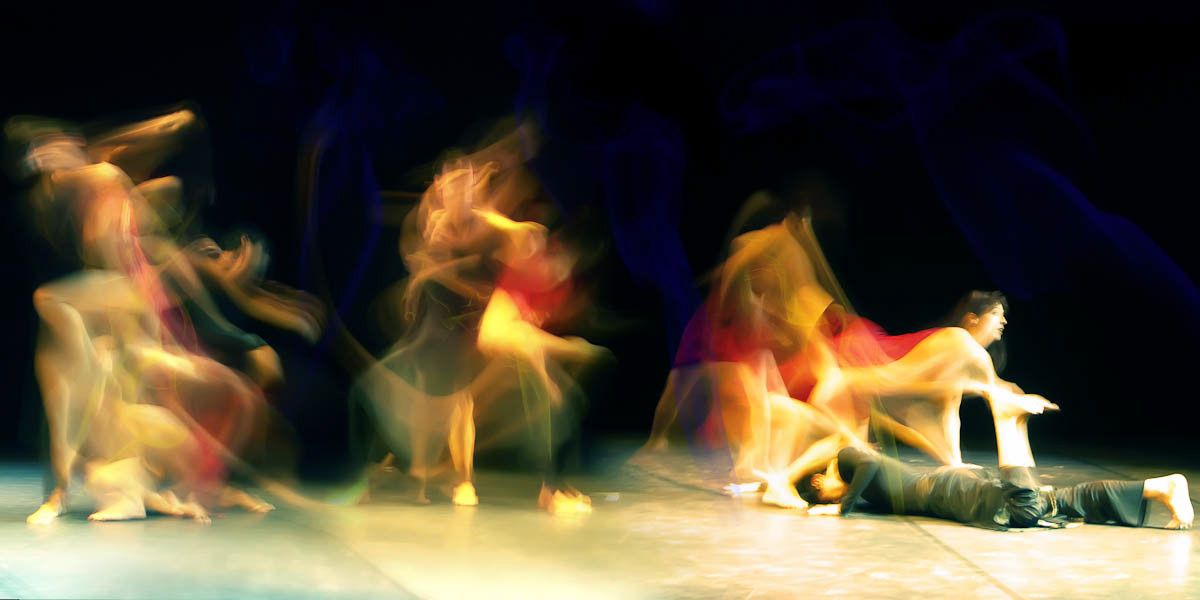 Wide stage with dancers in coloured costumes caught mid-movement, one lying on the floor.