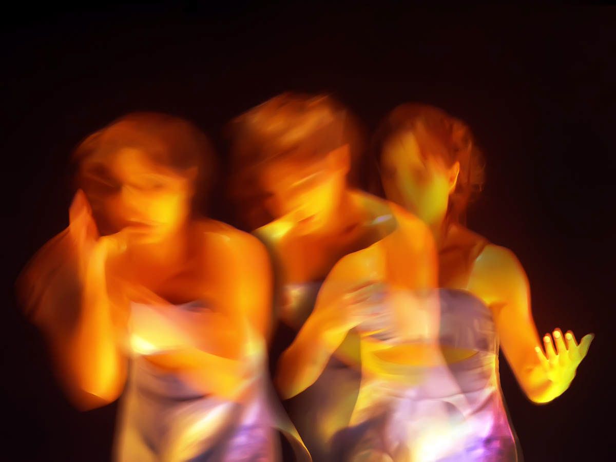 Three dancers glowing amber, faces half-hidden by blurred hands.