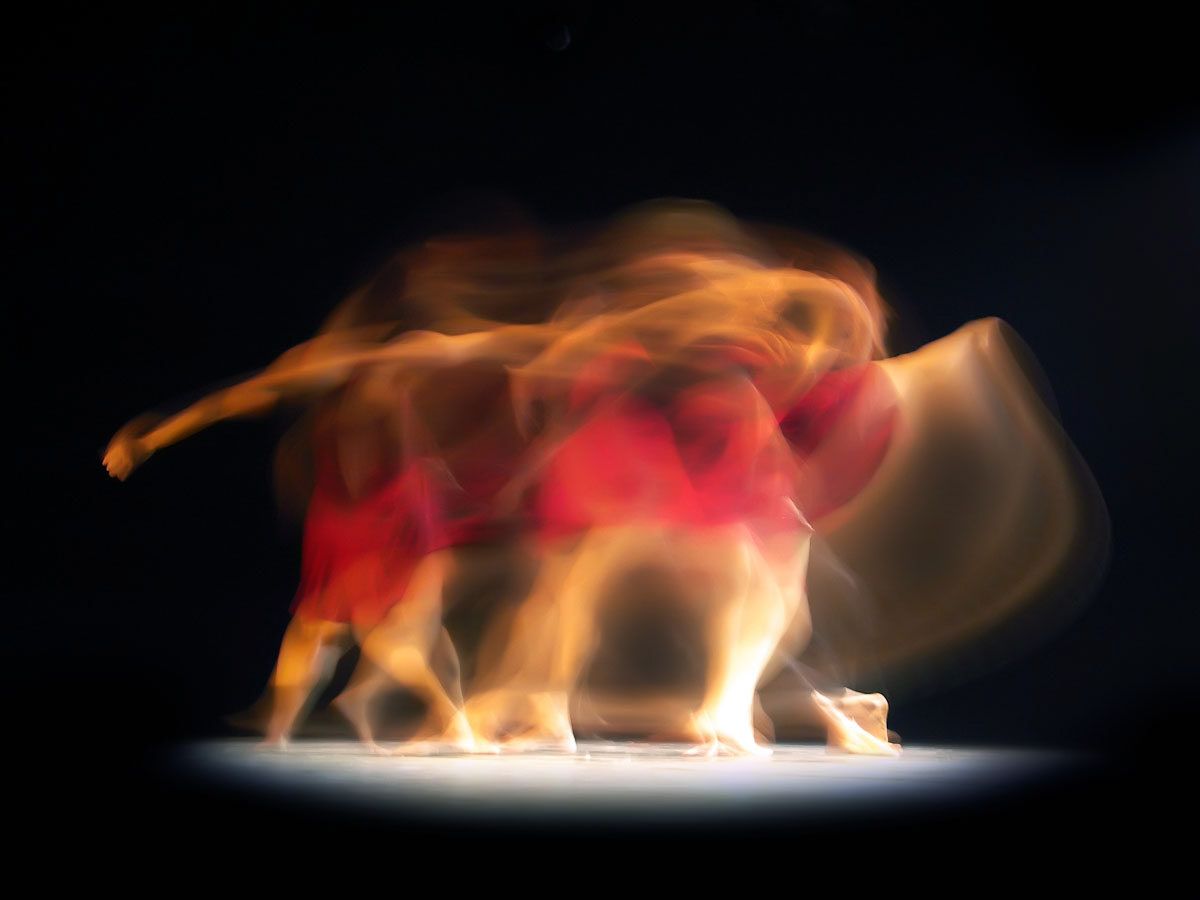 Dancers streaking sideways in long red and amber trails across a dark stage.