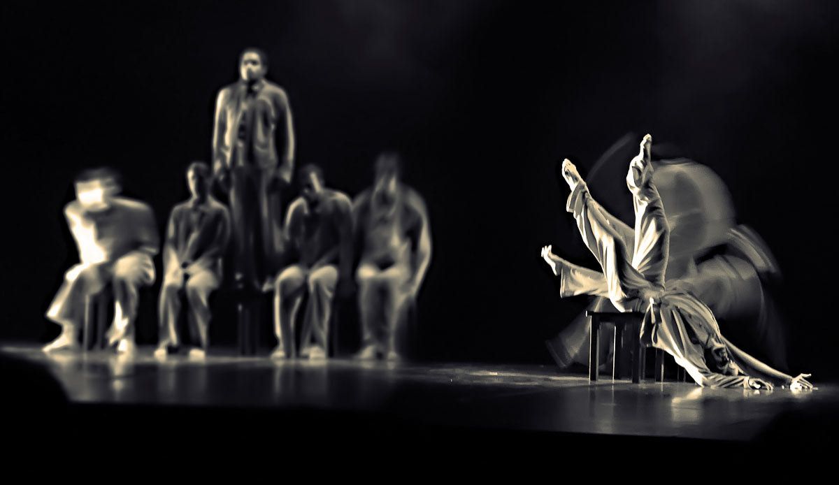 Dancers seated in a row on a dark stage, one standing behind, another bending low in a blur.