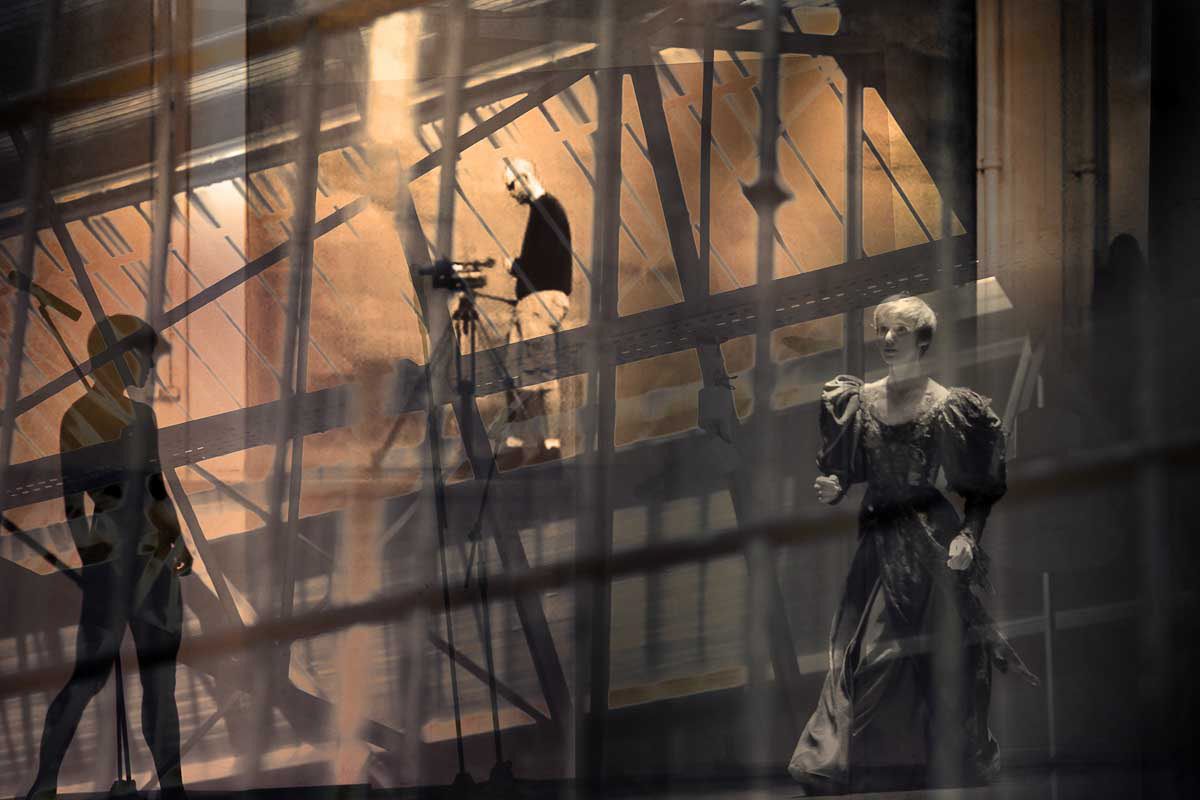 Figures, a mannequin and a camera on a tripod in a tangle of steel beams, warm light behind.