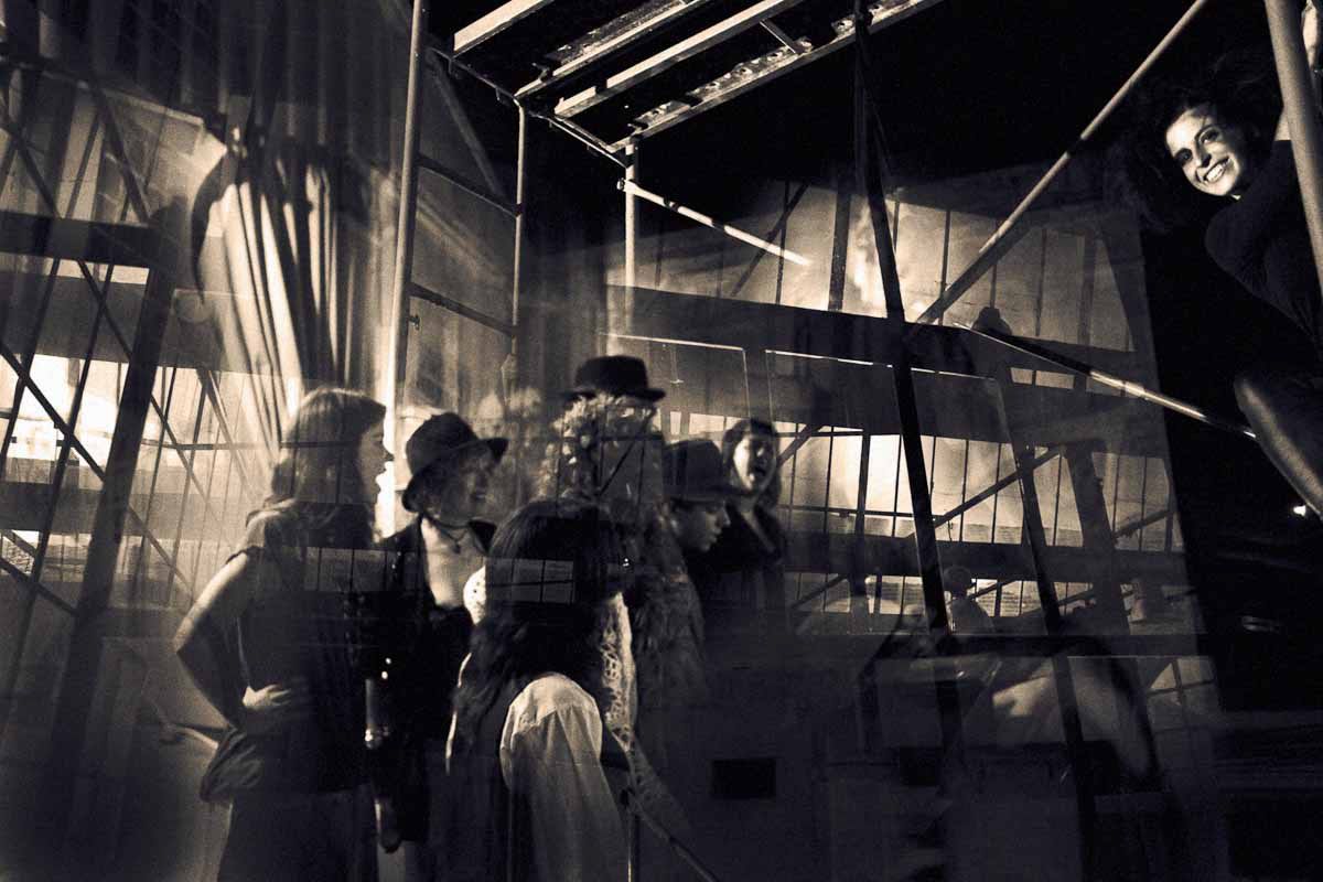 A group of figures under a steel structure, one woman smiling from the edge of the frame.