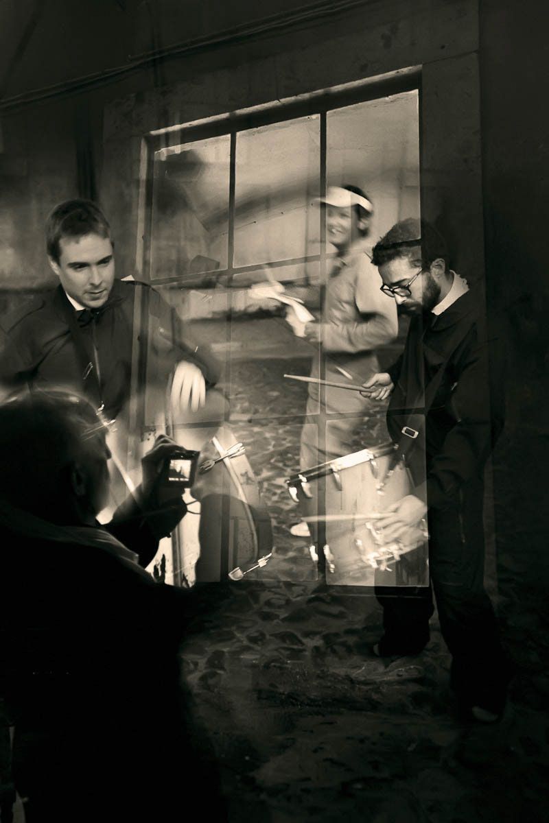Musicians with drums and a camera in a dim room by a tall window, in sepia.