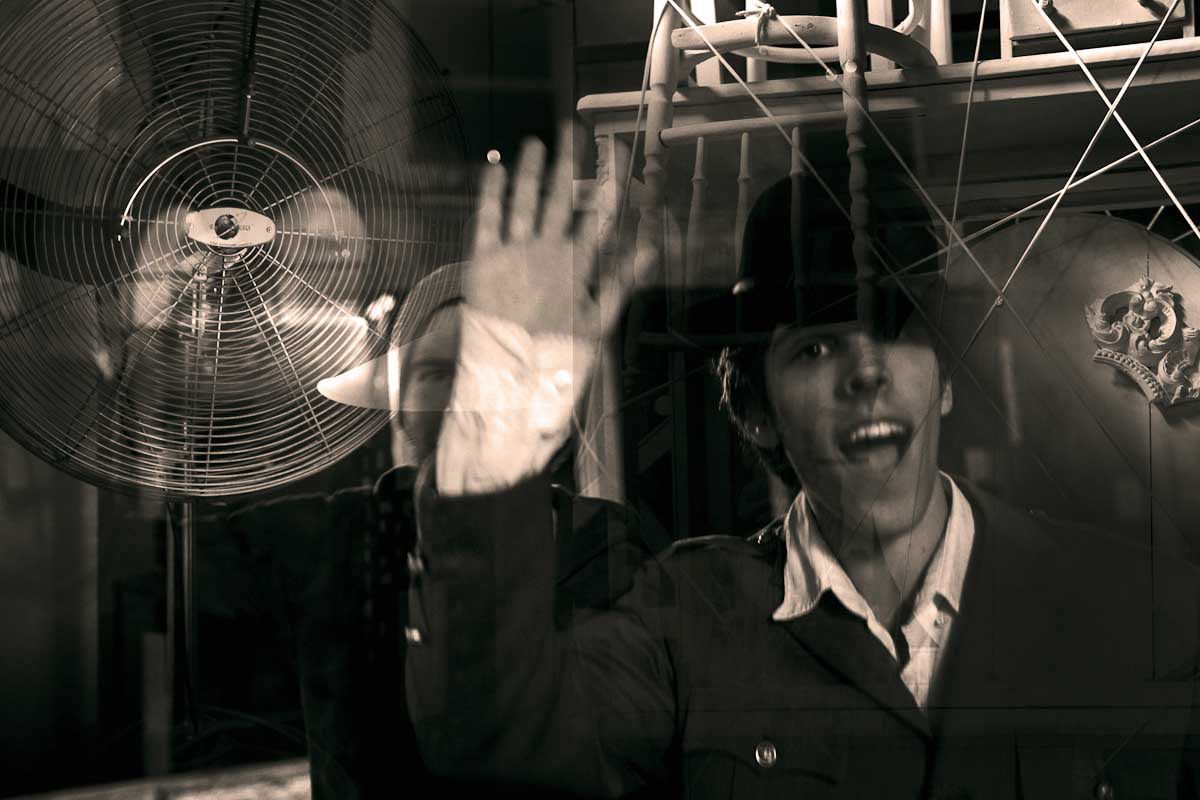 A young man in a bowler hat waving, behind a fan and a web of strings.