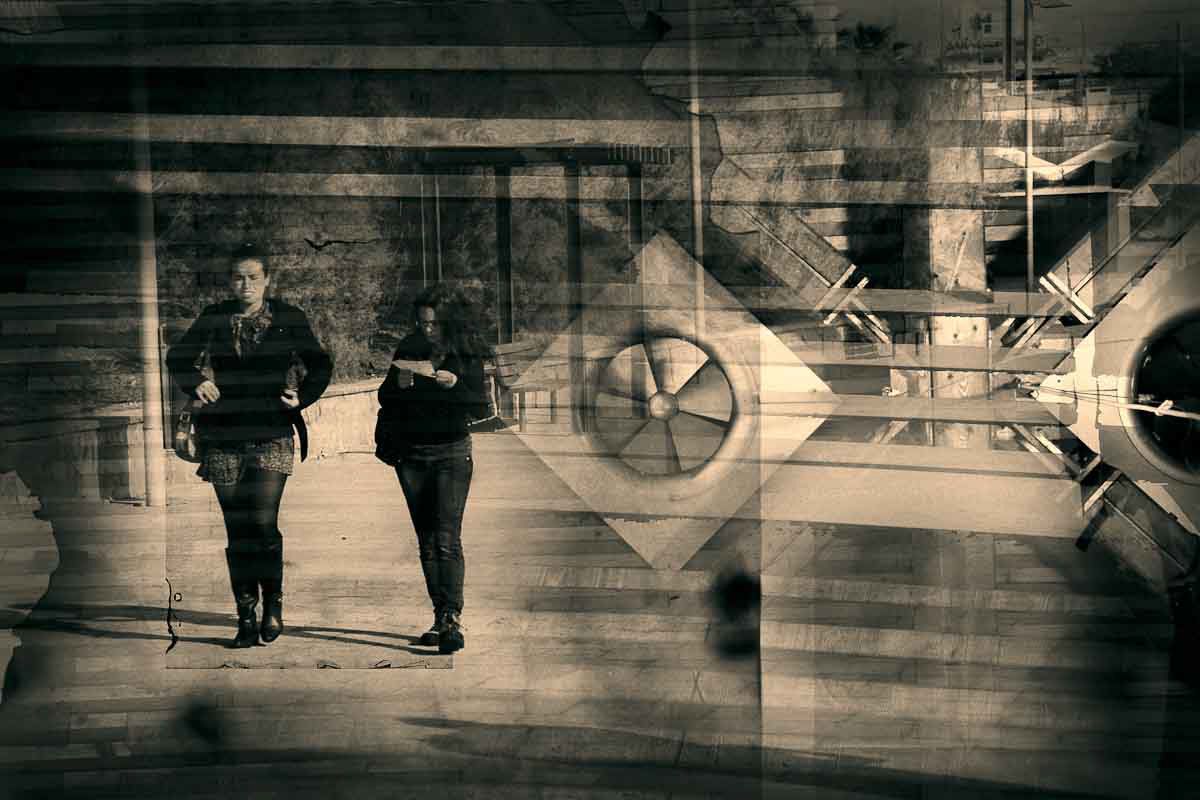 Two women standing in a pale, geometric space of beams and a large fan.
