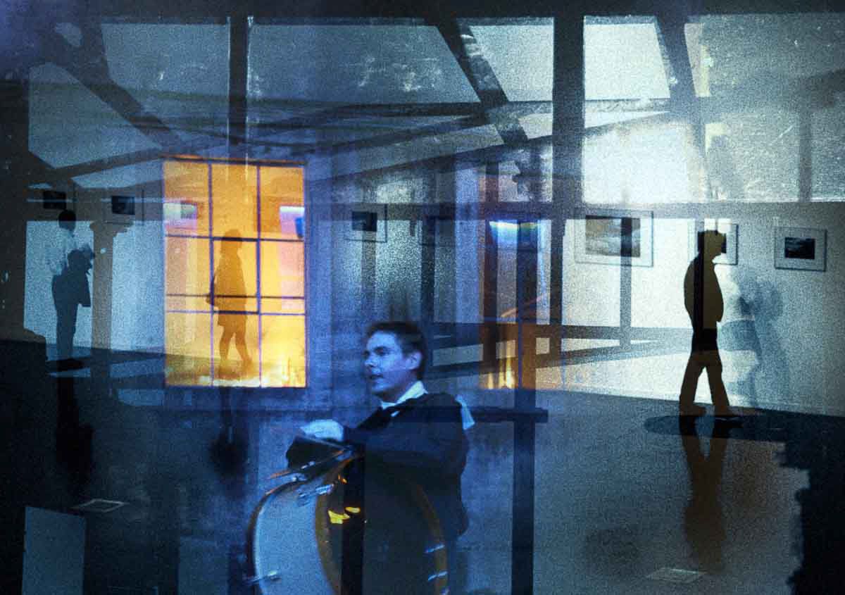A man with a drum in a blue gallery, a silhouette walking past lit frames.