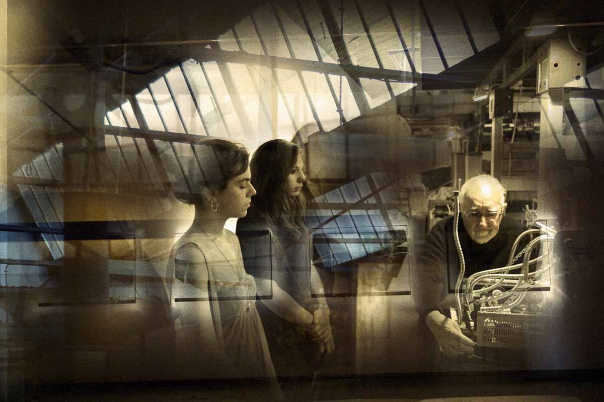 Two girls and an older man at a machine, under a glass roof in warm light.