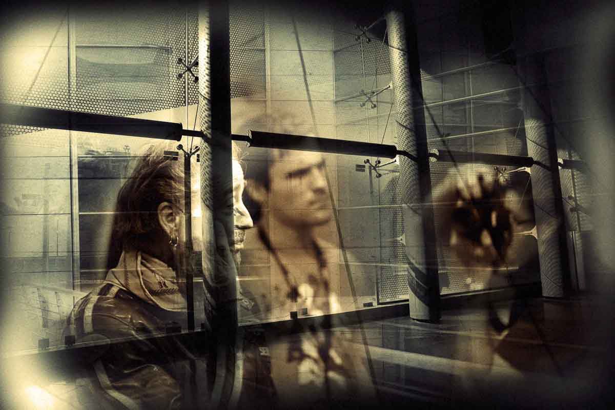 Two men behind a glass wall with columns, a blurred face on the right.