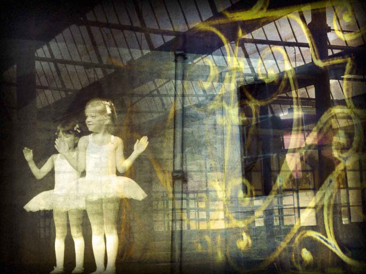 Two little ballerinas in white tutus beneath a glass roof and yellow scrollwork.