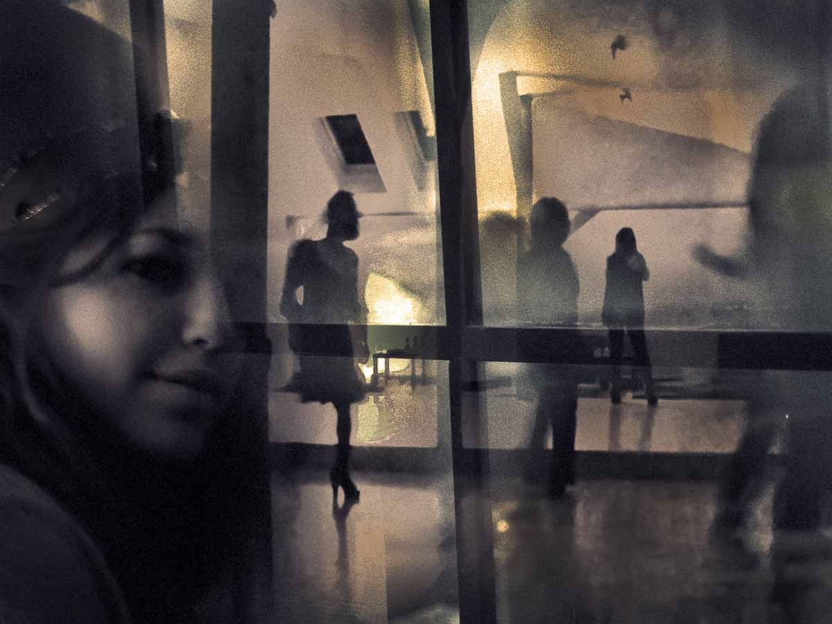A woman's face in the foreground, silhouettes walking in a hall behind glass.