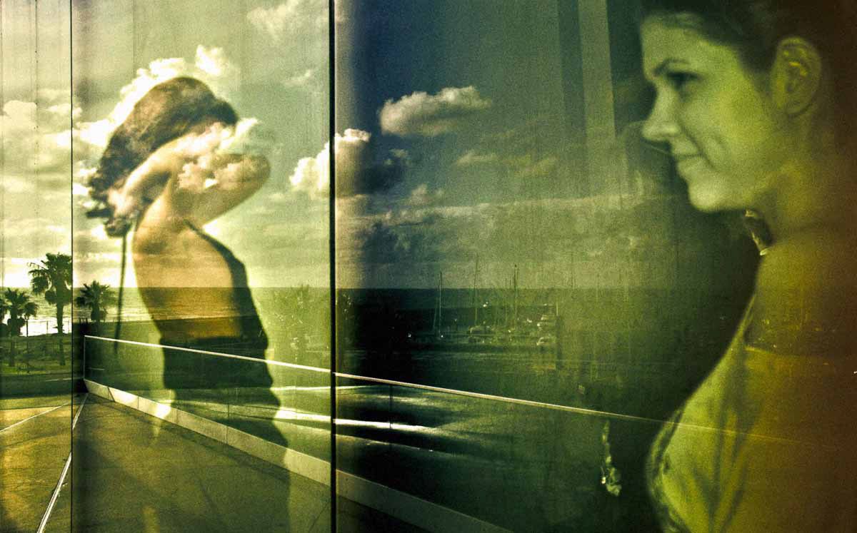 A woman looking at a view of palm trees and clouds reflected in glass, in green-gold tones.