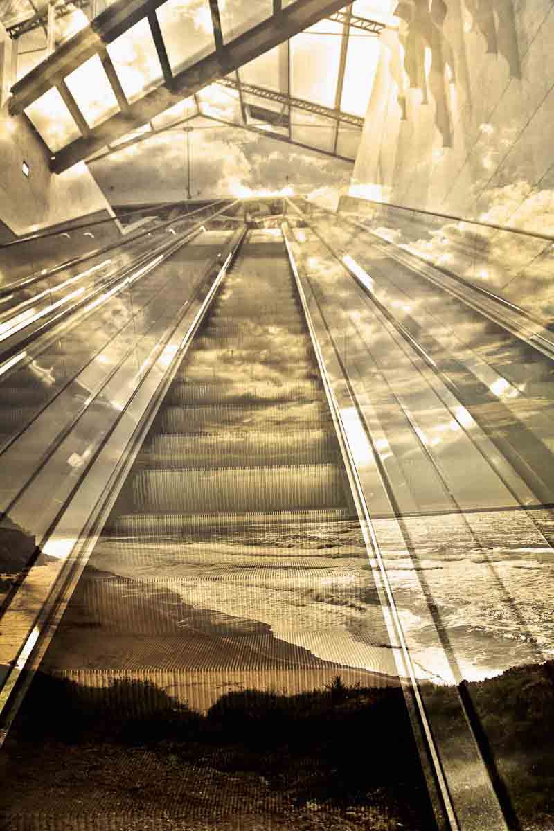 A long escalator rising under a glass roof, merging with a beach and the sea.