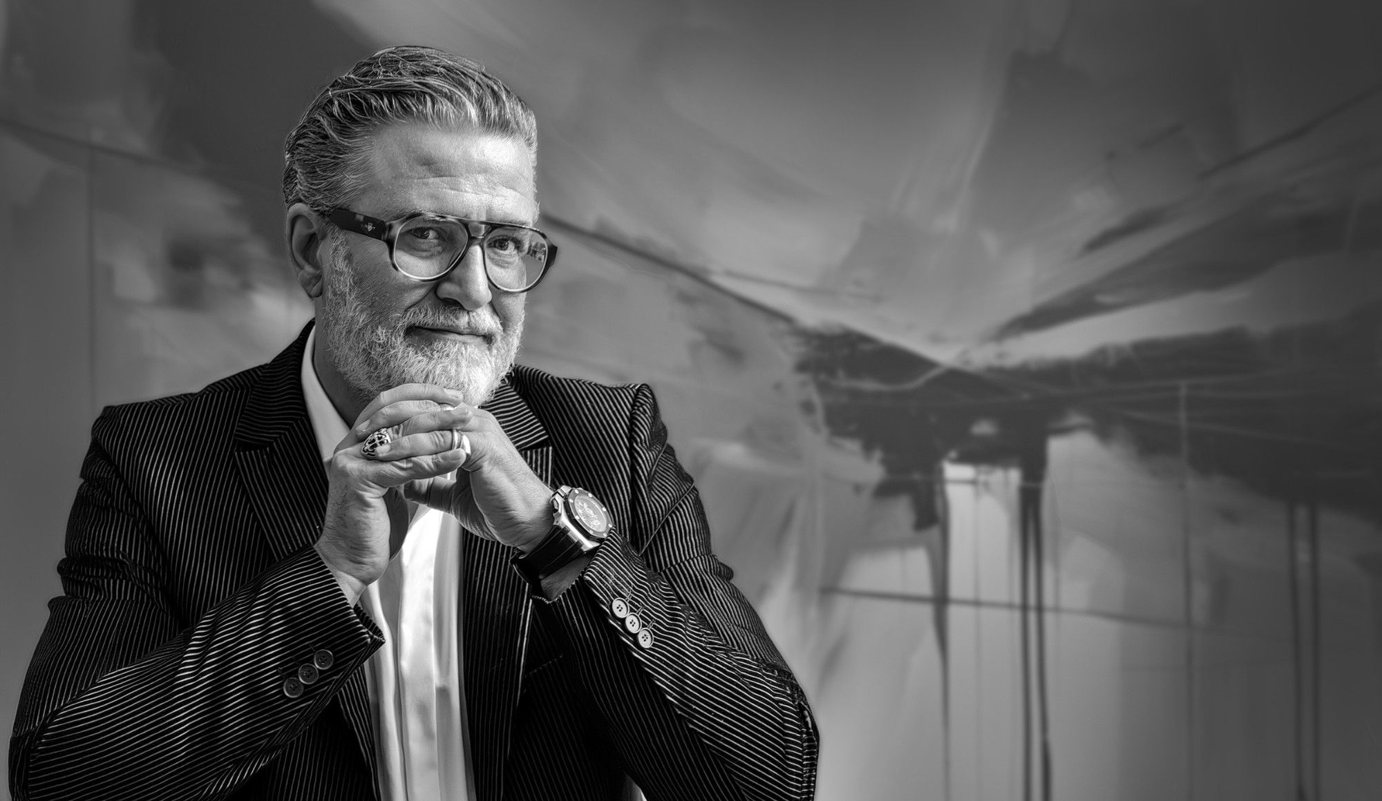 Black-and-white studio portrait of Dan St. Andrei, grey hair and beard, glasses, in a pinstripe jacket, hands clasped near his chin, in front of an abstract gestural backdrop.