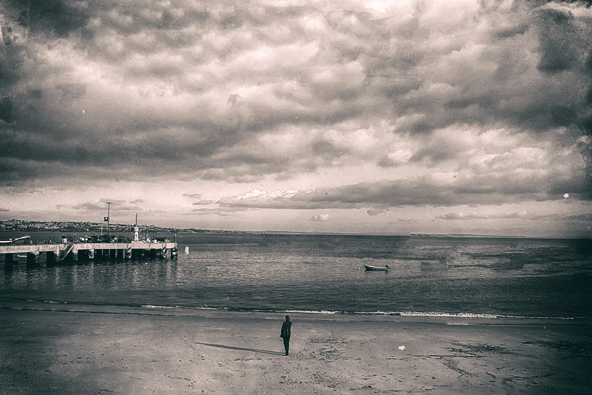 A lone figure on a wide beach at low tide under a heavy clouded sky, a pier in the distance, in sepia.