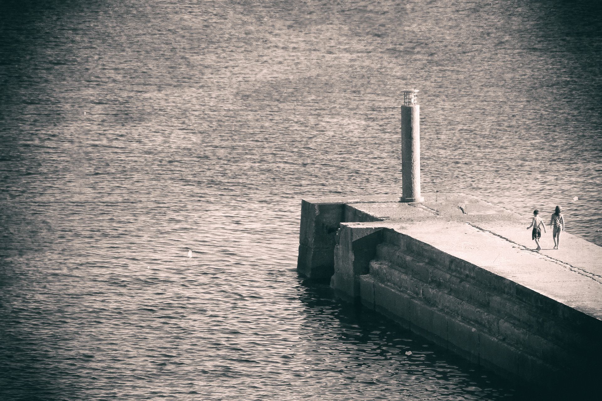 A stone jetty with a tall column reaching into a rippled sea, two small figures walking along it.