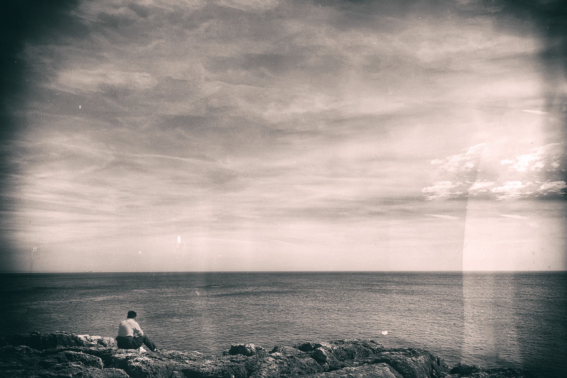 A person sitting on dark rocks facing a calm open sea under a pale sky.