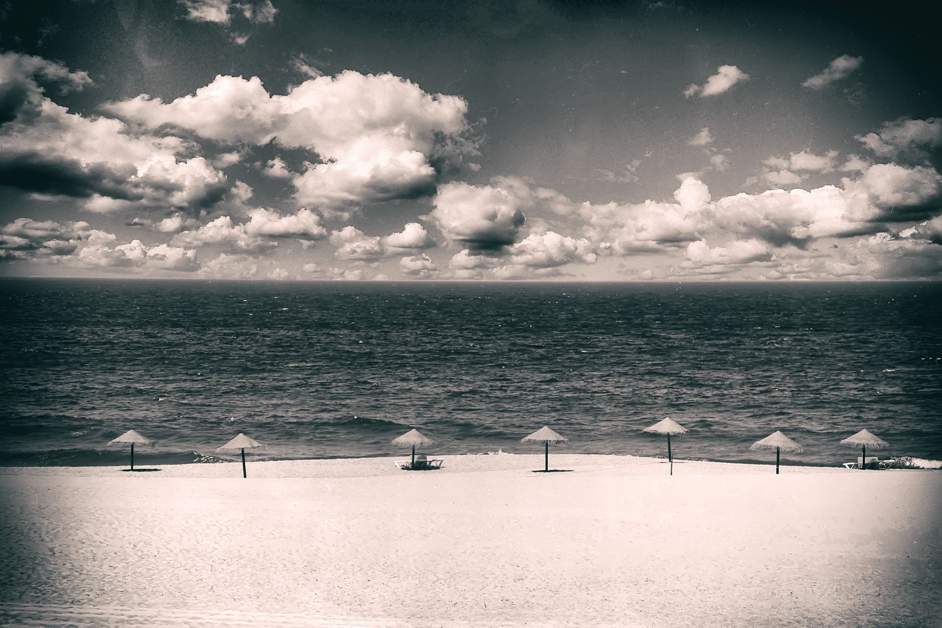A row of straw parasols on an empty white beach before a dark sea and bright clouds.
