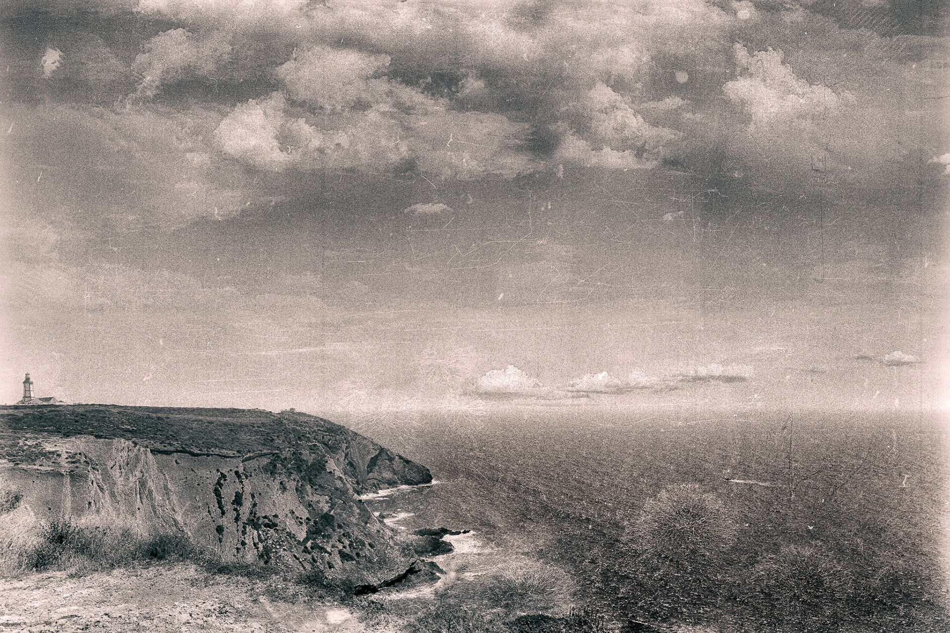 A rugged headland with a small lighthouse above the ocean, seen from a grassy slope.