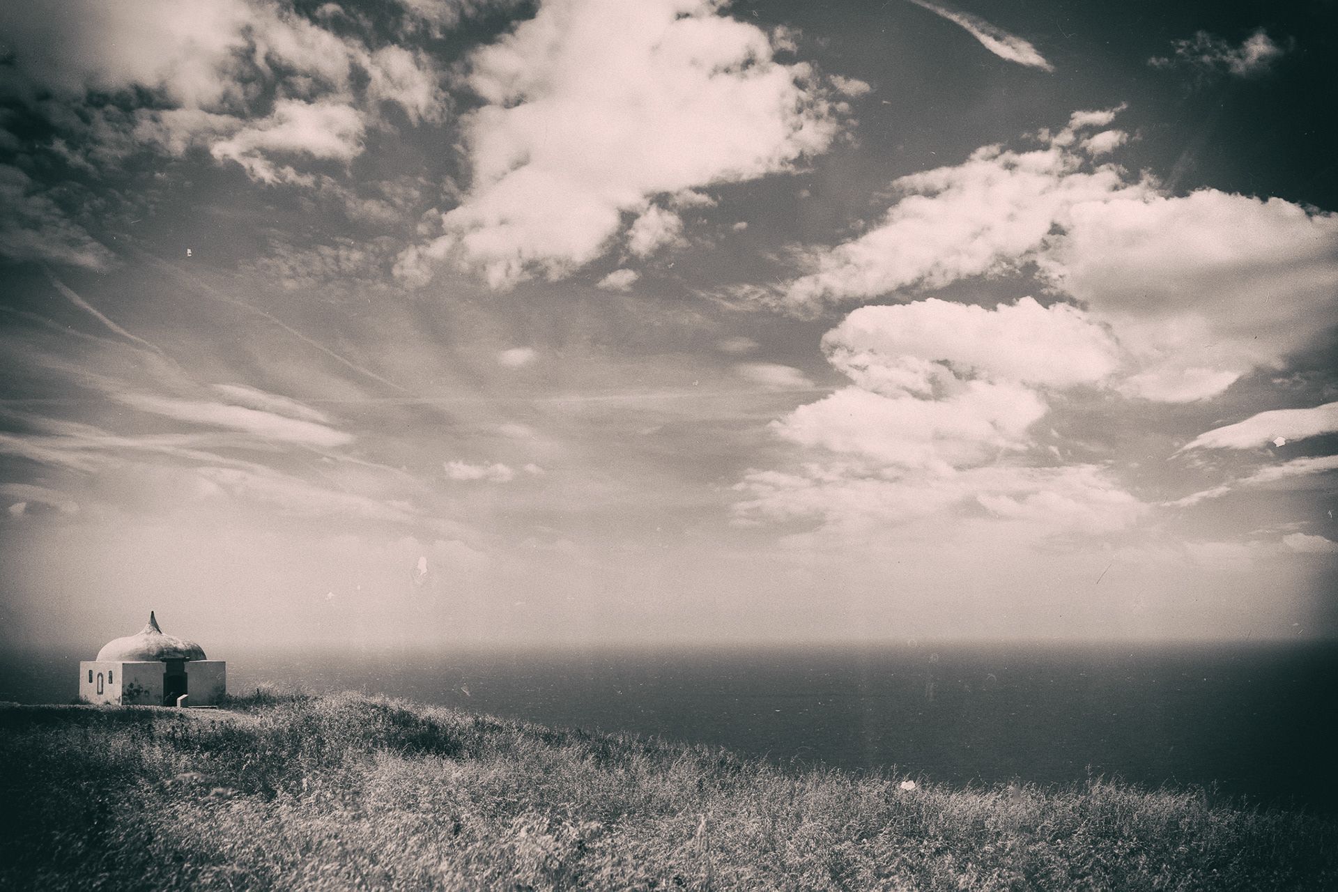 A small white chapel on a grassy cliff above the Atlantic, under a wide sky of clouds.