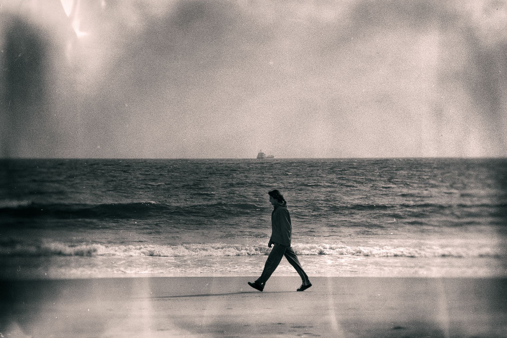 A man in a long coat walking along the water's edge, a ship far out on the horizon.