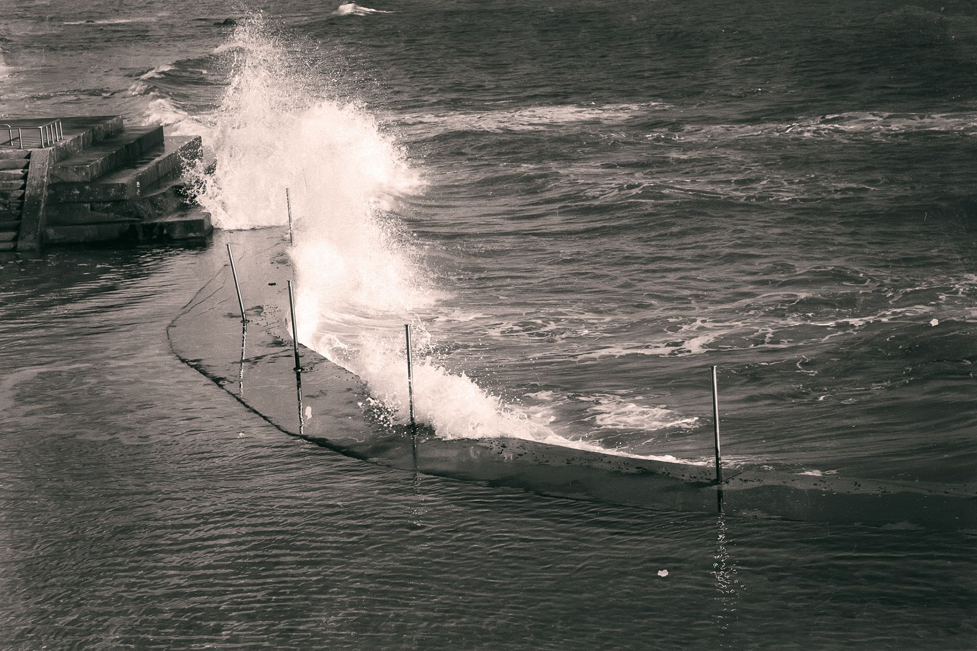 A wave breaking in white spray over a low sea wall with thin posts.
