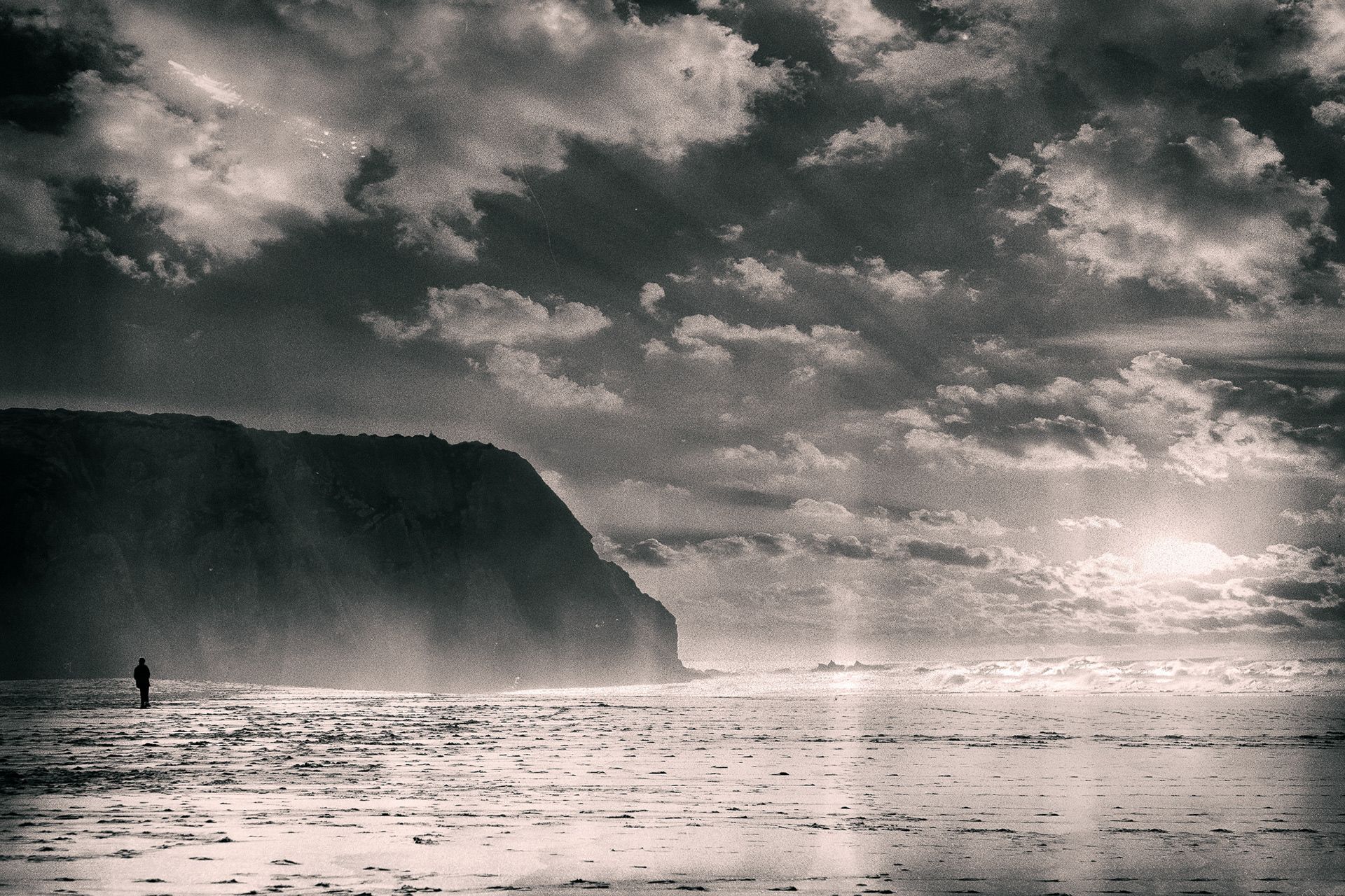 A tiny figure on a beach below a dark cliff, under dramatic clouds and hazy light.