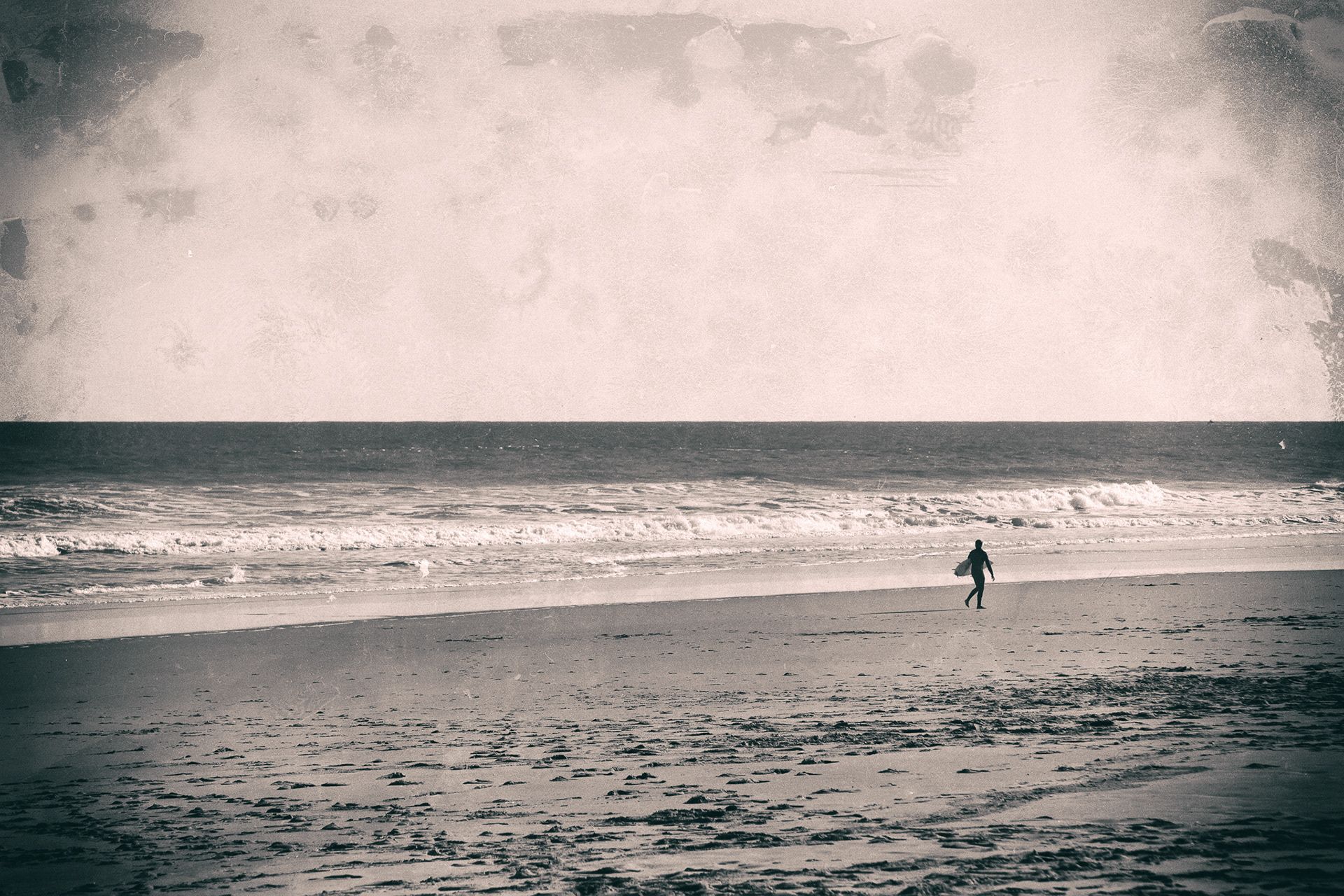 A surfer running across a wide beach toward the waves under a pale sky.