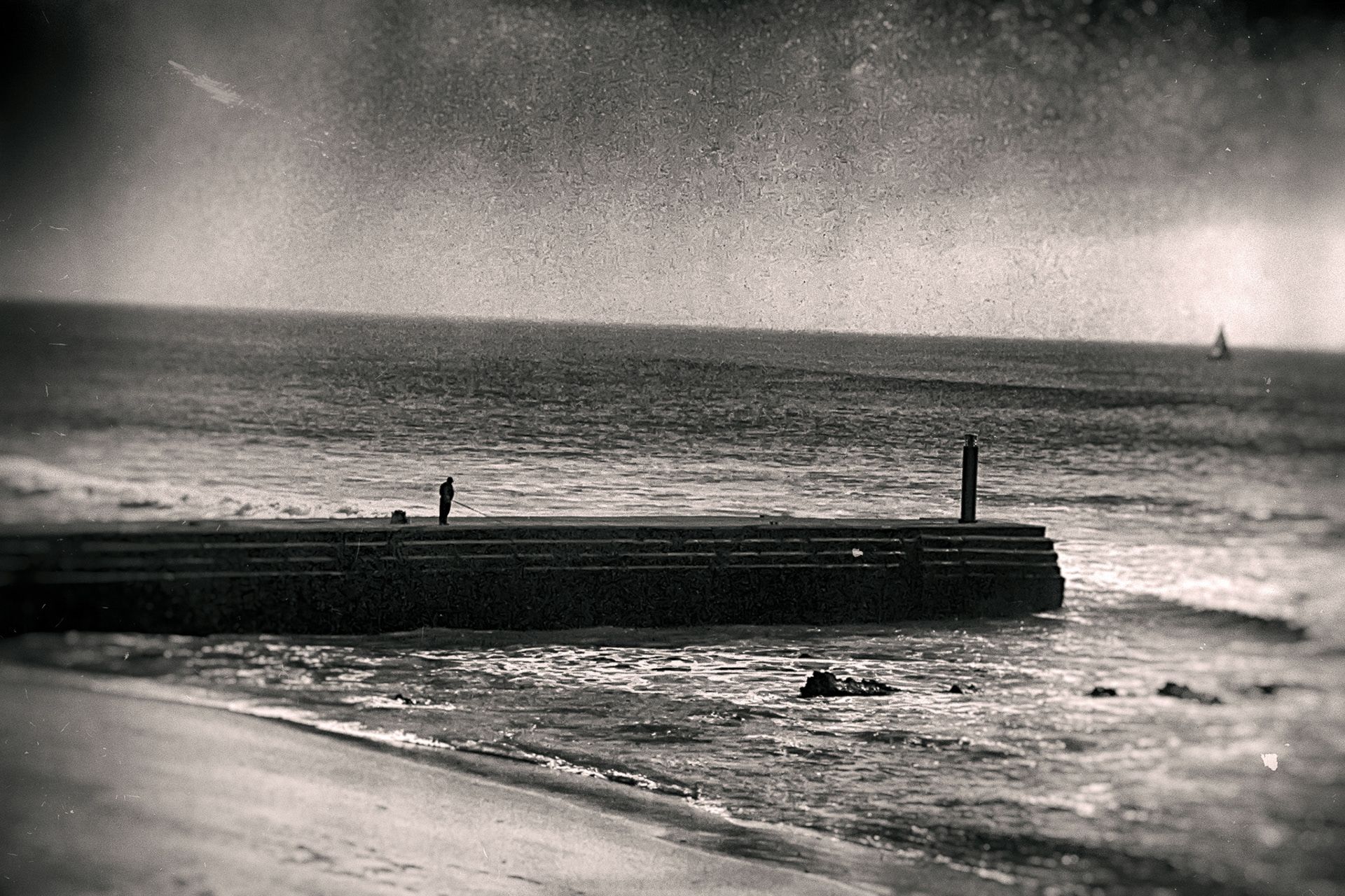 A long jetty running into the sea with two standing figures, a sailboat on the horizon.