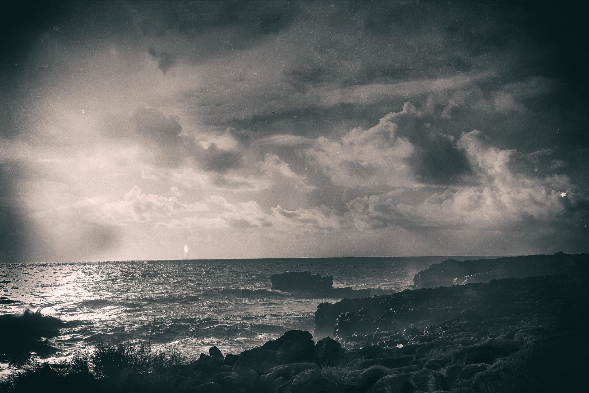 A rocky shoreline under a dark, clouded sky, light glinting on the water.