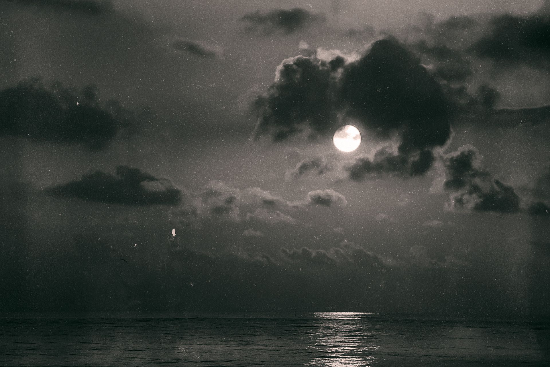 A full moon breaking through dark clouds above the sea, its light reflected on the water.