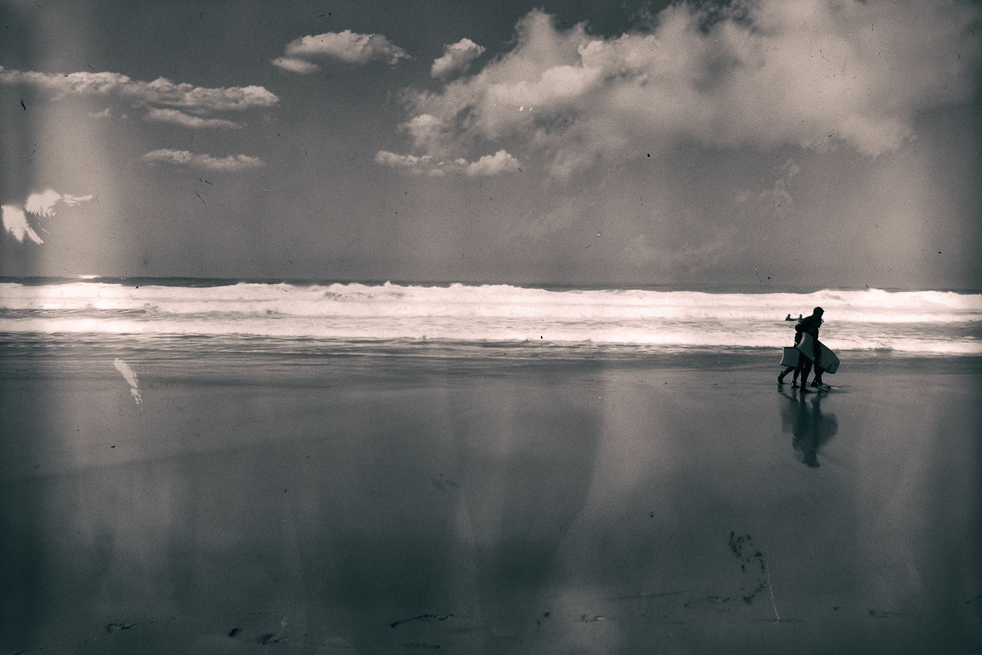 Two figures walking along wet sand that mirrors them, waves and clouds behind.