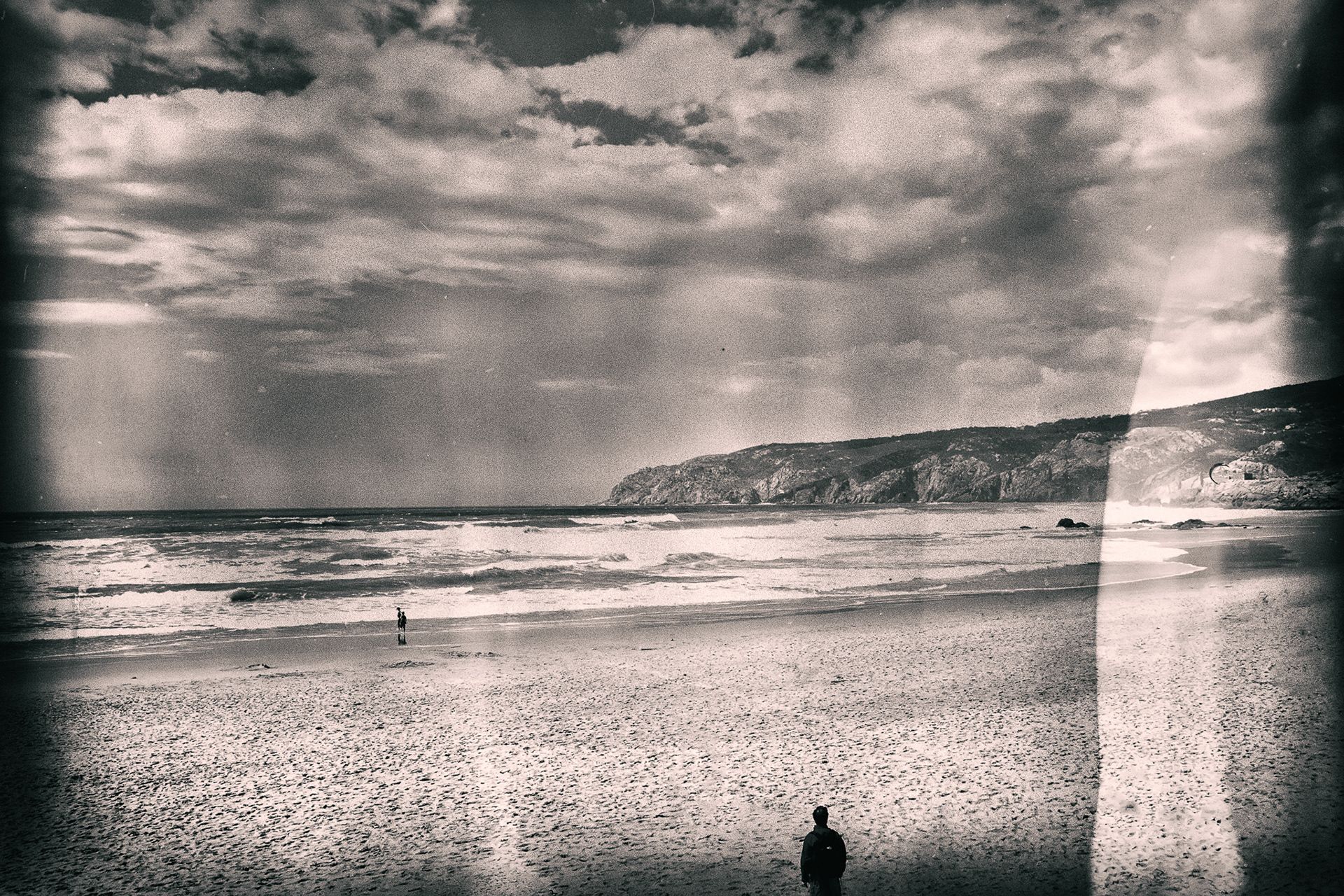 A vast beach under a stormy sky with rays of light, a lone figure in the foreground.