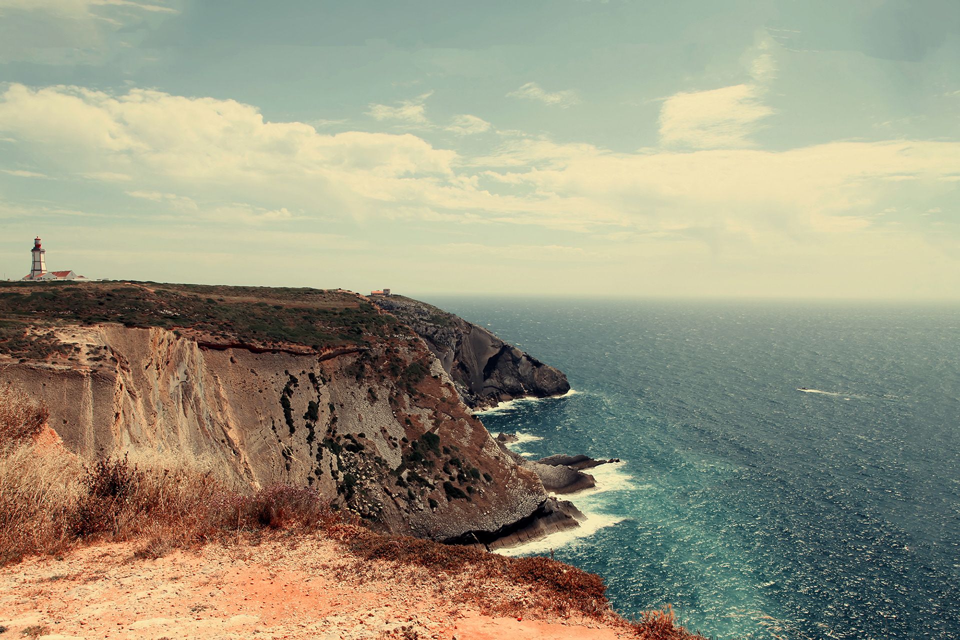 Ochre cliffs and a lighthouse above a turquoise sea, in faded colour.