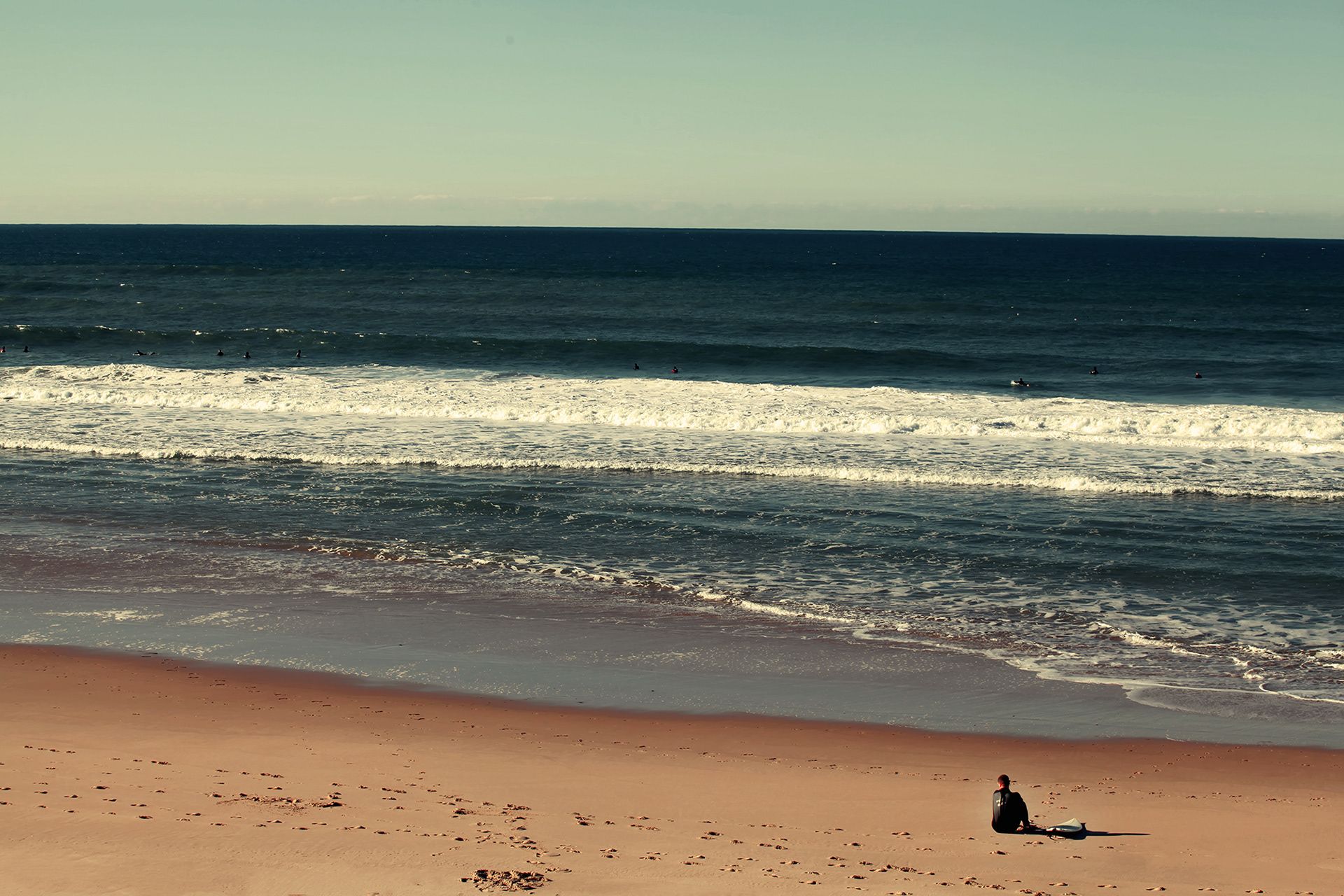A person sitting on a reddish beach before white breaking waves and a deep blue sea, in colour.